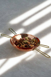 Copper tea strainer with brass handles holding dried herbs and tea leaves on a rice paper background with gentle light and intricate shadows