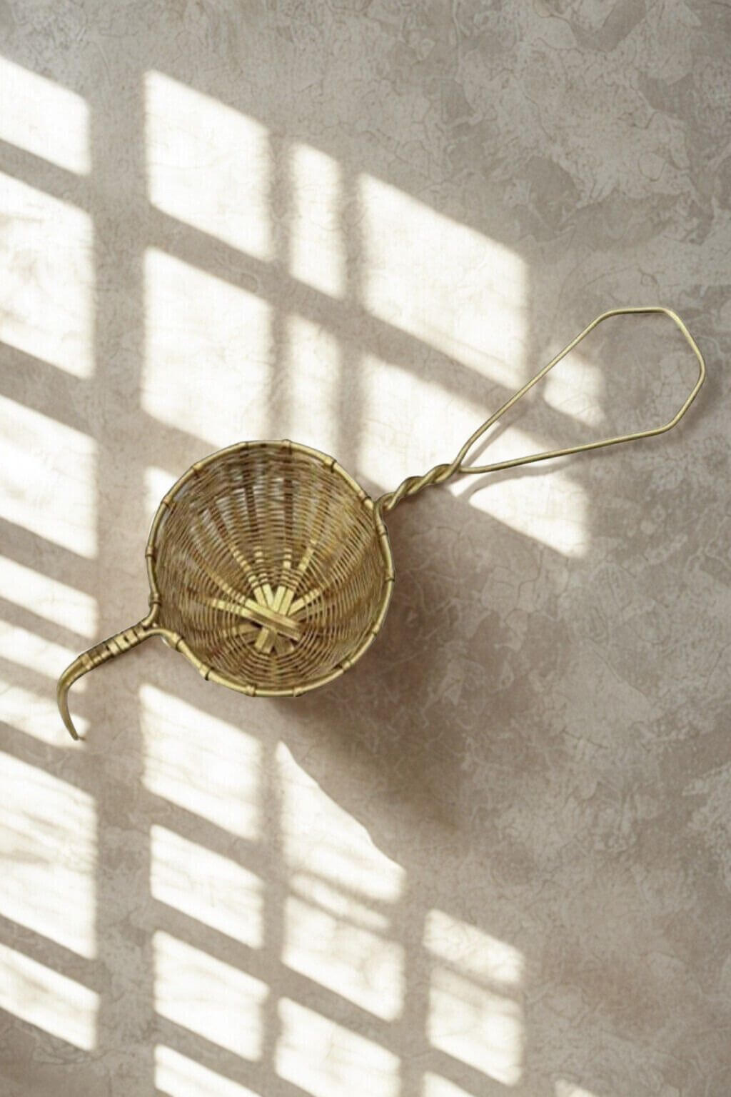 Close-up of woven raw brass tea infuser on a textured stone surface with sunlight casting shadows.