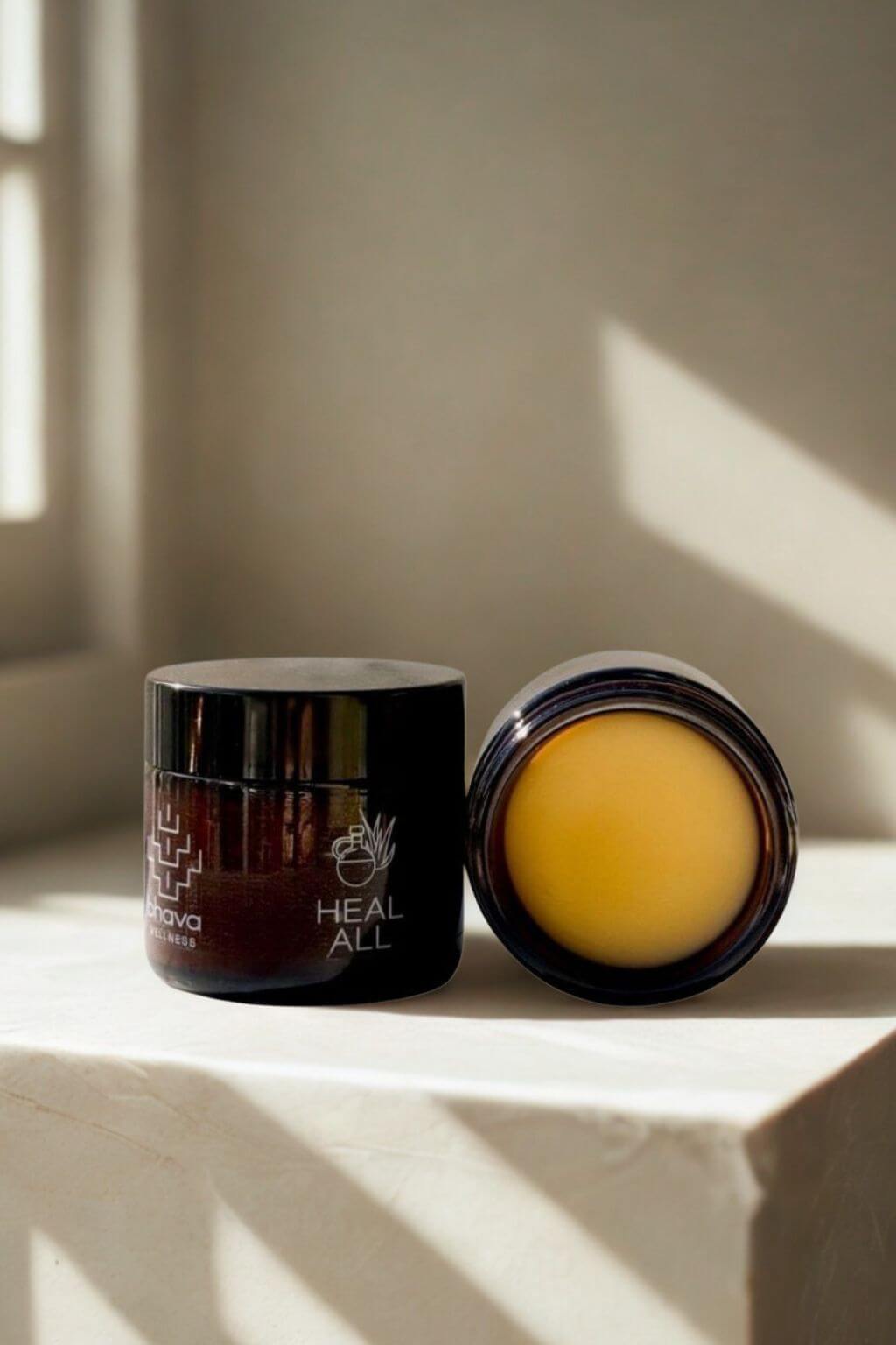 Two jars of Bhava Wellenss Heal All Herbal Salve in a light filled space with warm shadows from the paned window.
