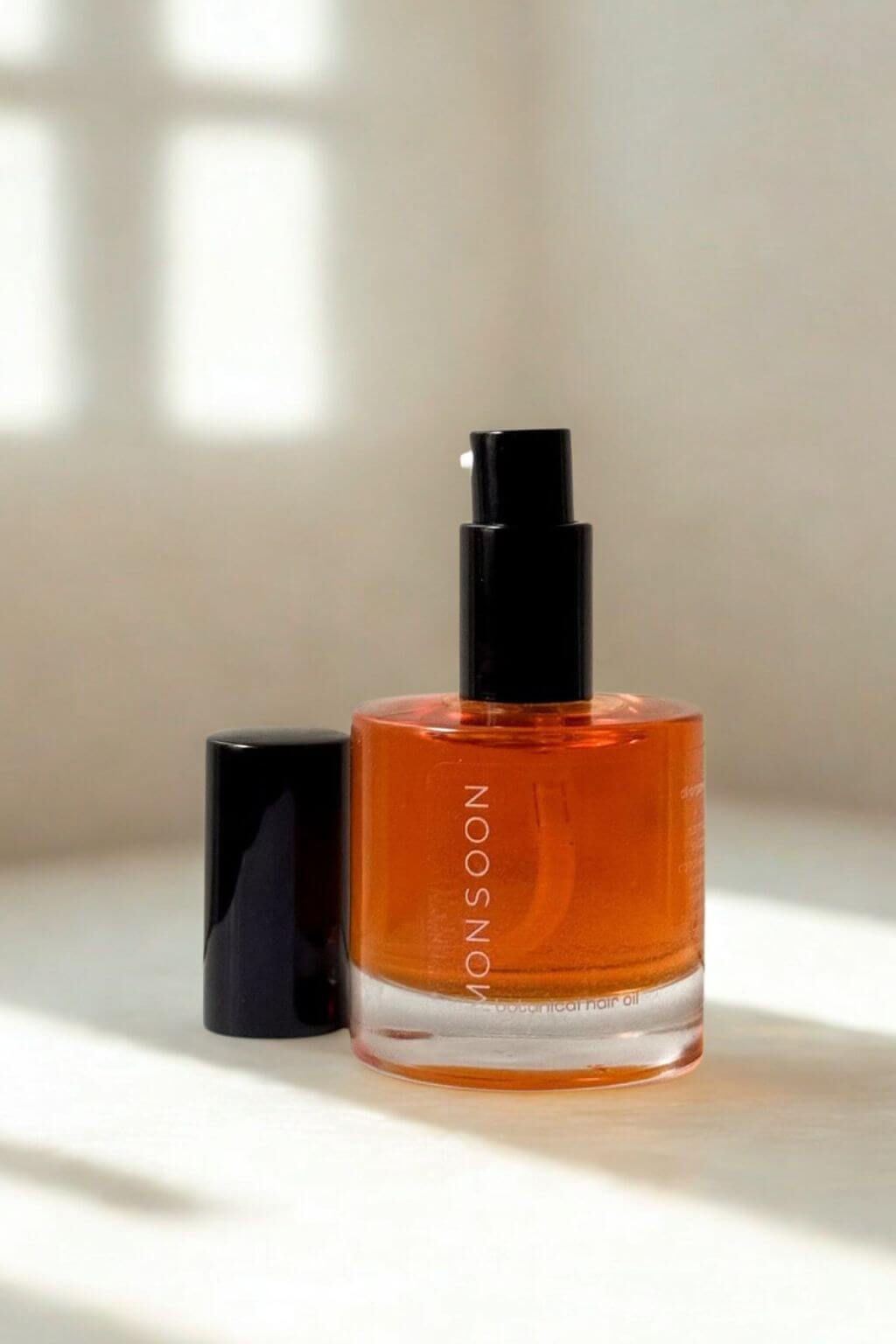 Serum bottle with Monsoon Botanical Hair Oil alongside black cap on a light surface, with shadows from the window against a soft white ackground