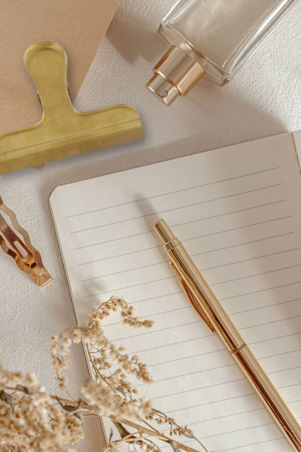 A gold pen resting on an open lined notebook, styled with dried flowers, a gold clip, and a clear glass perfume bottle on a textured beige surface.