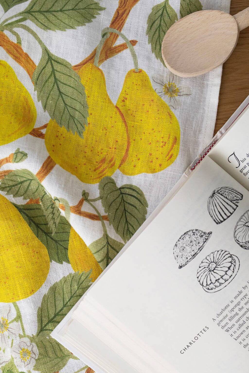 Pear Orchard Linen Tea Towel with cookbook and wooden spoon, styled for a cozy kitchen scene.