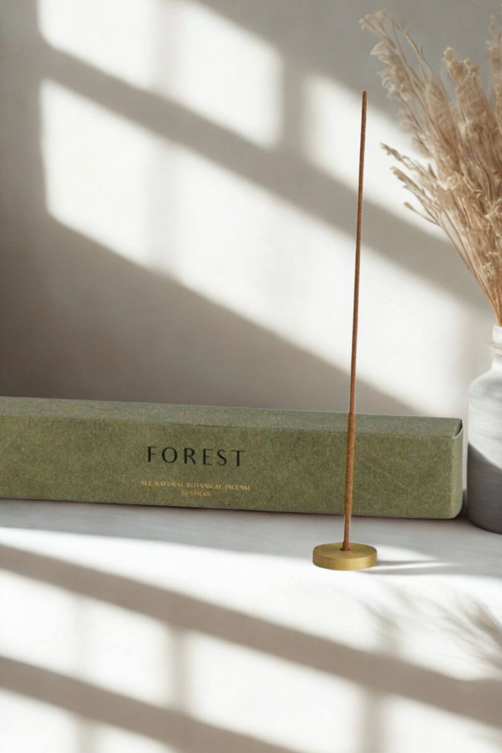 Minimalist brass incense holder with a lit cedarwood incense stick, displayed beside a green "Forest" box on a sunlit surface with pampas grass in a ceramic vase.