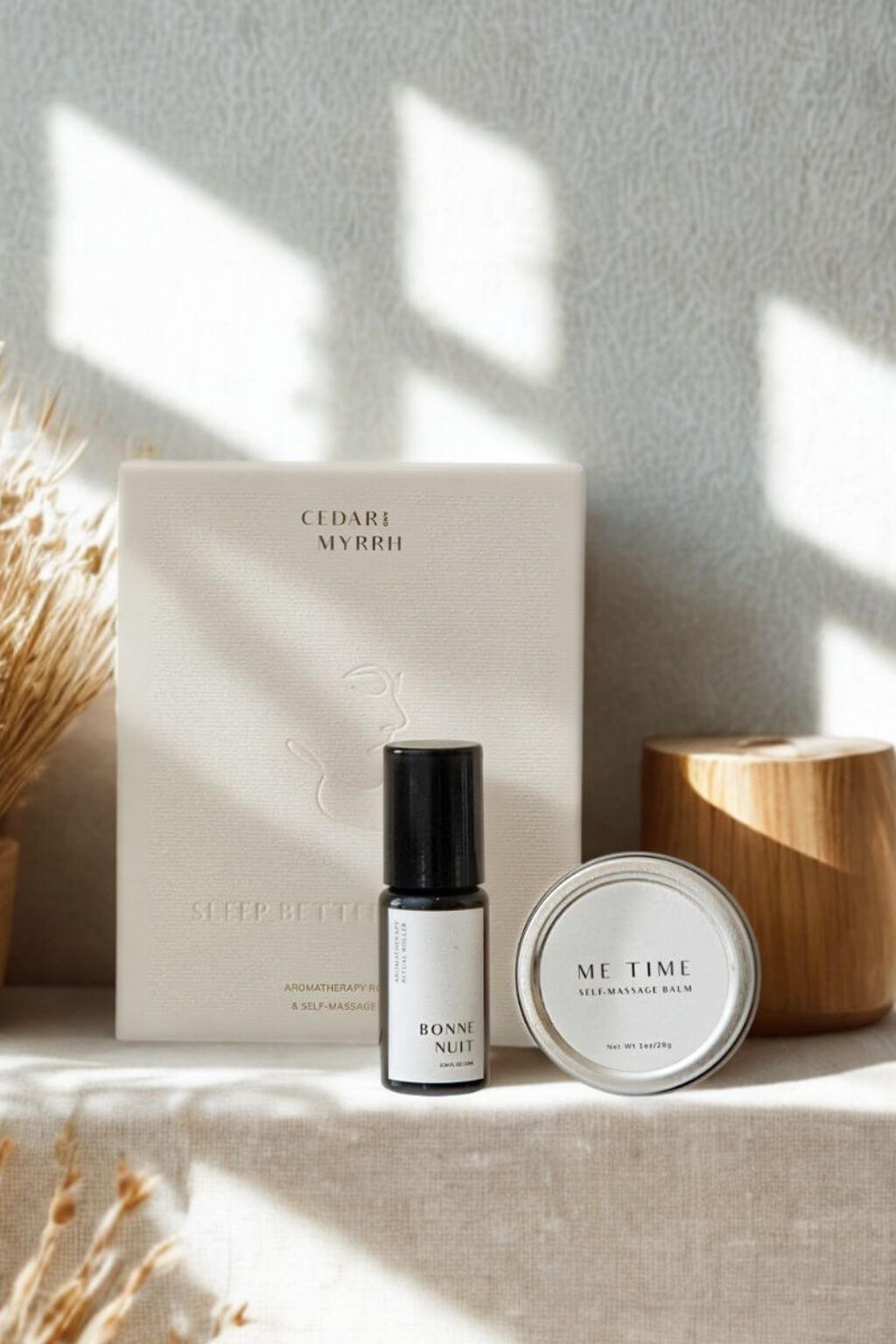 Cedar + Myrrh roller bottle of Bonne Nuit, and Me Time Balm with embossed gift box on a textured surface with natural light.