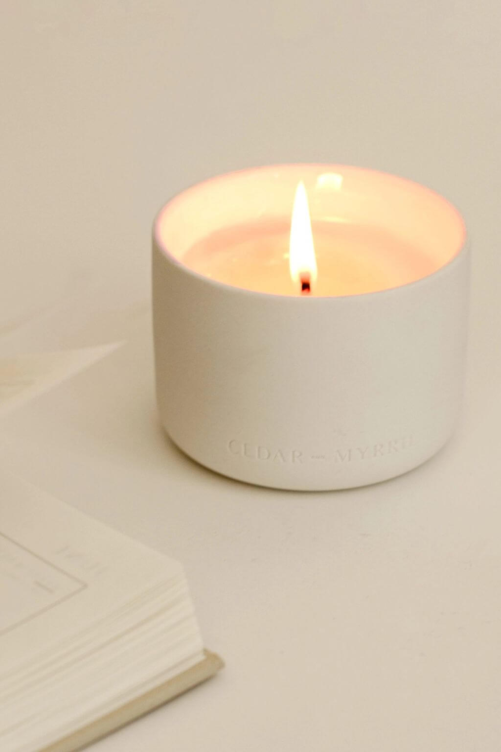 A lit Mantra Wellness Ritual Candle in a matte ceramic vessel, styled beside an open book for a serene moment of relaxation.