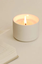 A lit Mantra Wellness Ritual Candle in a matte ceramic vessel, styled beside an open book for a serene moment of relaxation.