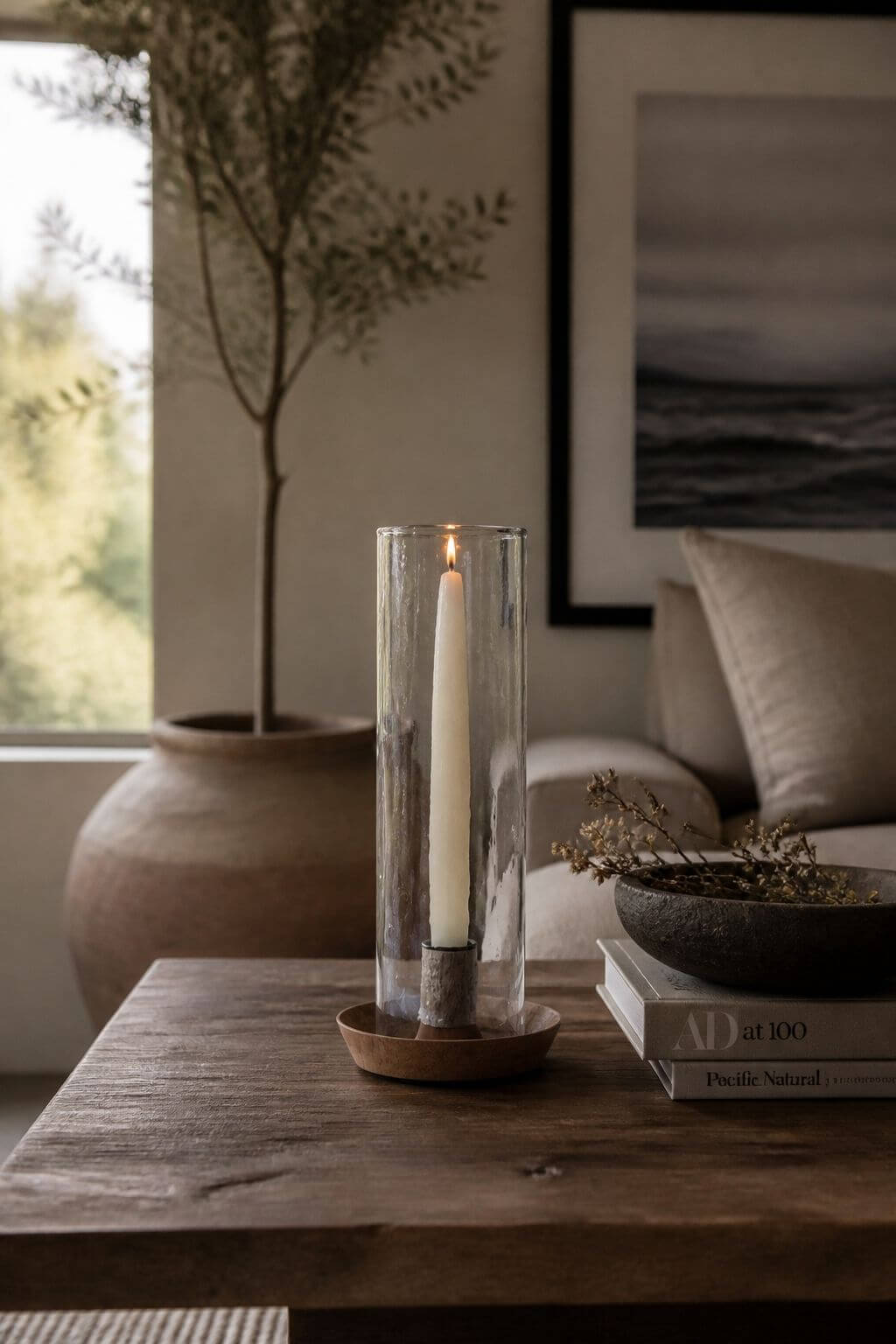 A tall clear glass cylinder candle holder with a lit white taper candle, set on a wooden base on a rustic wooden table in a cozy living room setting.