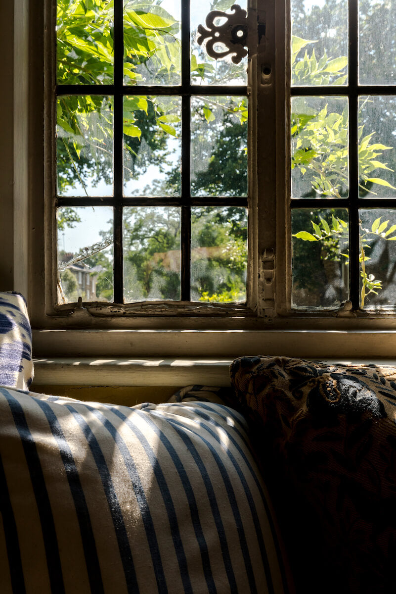 A window seat with blue and white striped pillows, with a view of vibrant green foliage outside a pane glass window.