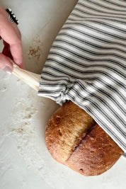 Person pulling drawstring closure on a ticking stripe bread bag filled with a loaf of homemade bread