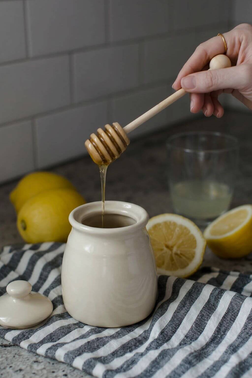 A hand holds a wooden honey dipper above a small cream-colored ceramic jar, with honey dripping into the jar. Lemons, a striped cloth, and a glass are visible on a kitchen counter against a gray subway tile backsplash.