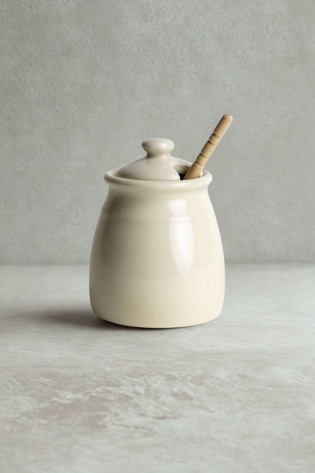 A cream-colored ceramic honey pot with a matching lid and a wooden dipper spoon, set against a neutral gray textured background.
