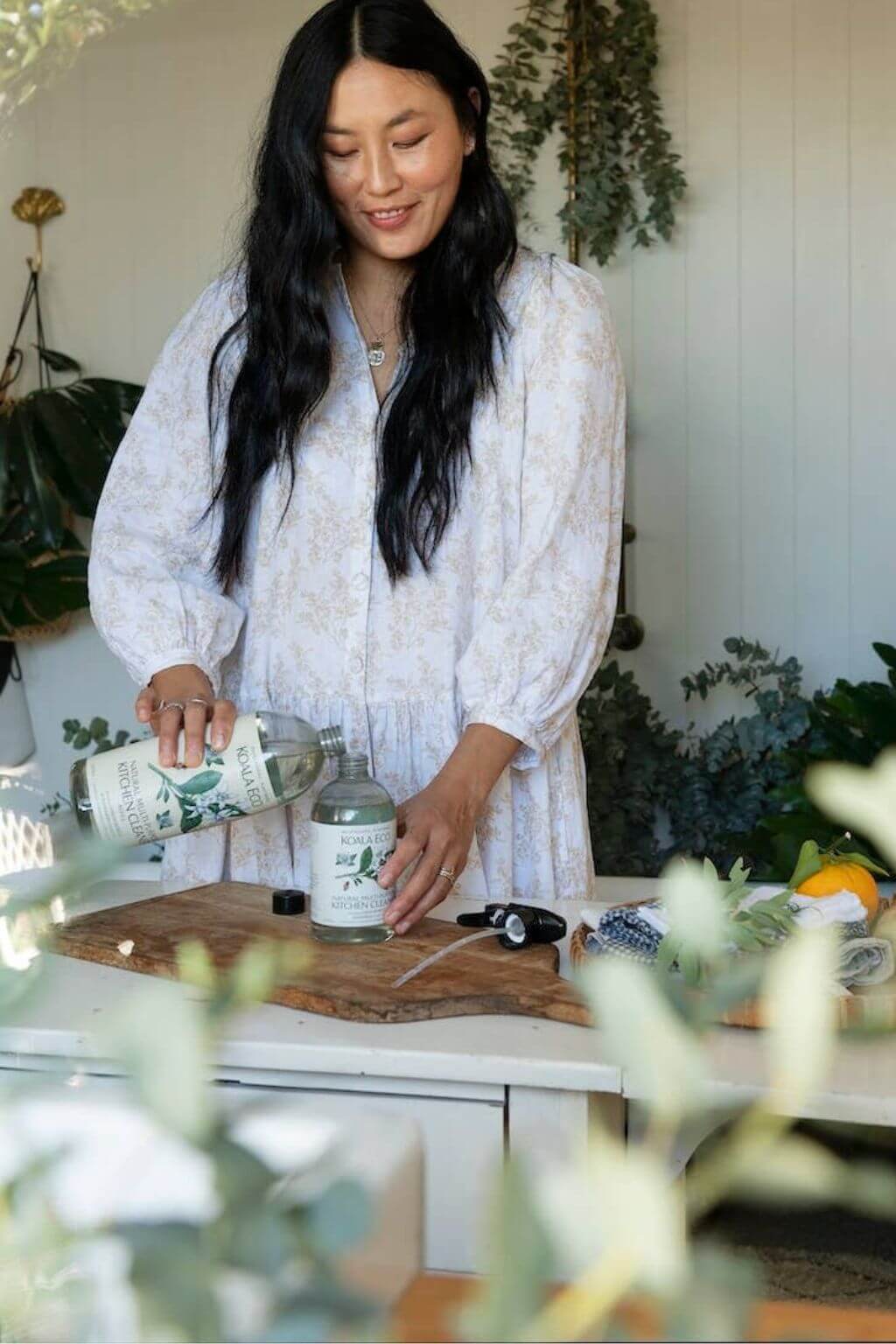 Woman refilling her Koala Eco Multi-Purpose Kitchen Cleaner on a wooden cutting board with plants in the background.
