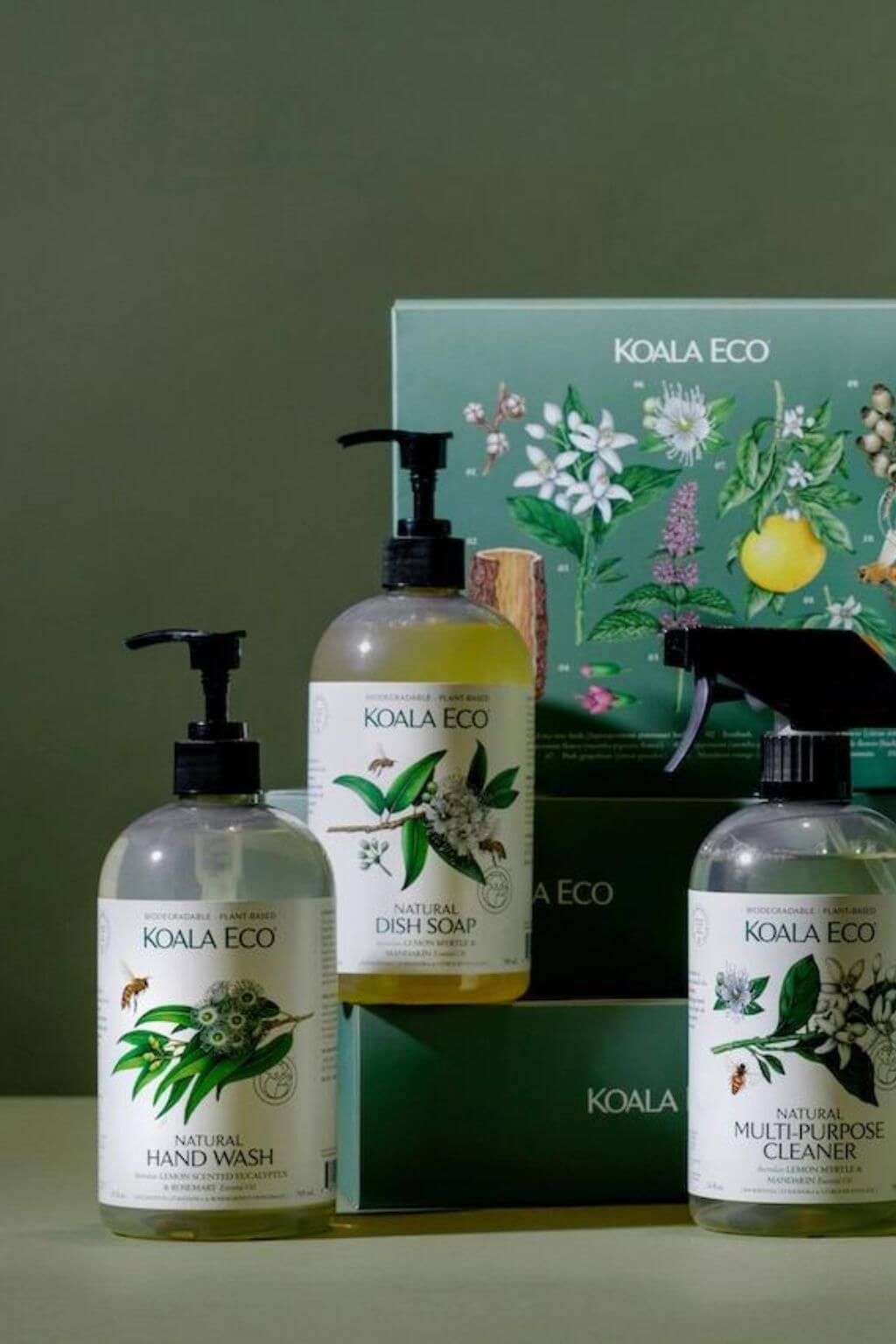 A set of Koala Eco Natural Cleaning Products, including hand wash, dish soap, and multi-purpose cleaner, displayed in front of a dark green gift box with botanical drawings.