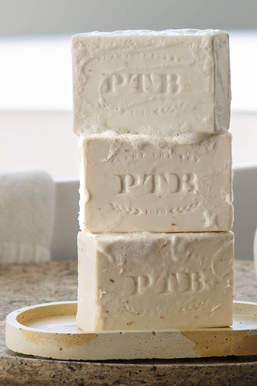 Three stacked rectangular bars of PTB soap on a round ceramic dish, photographed in a softly lit bathroom-like setting with a towel blurred in the background.