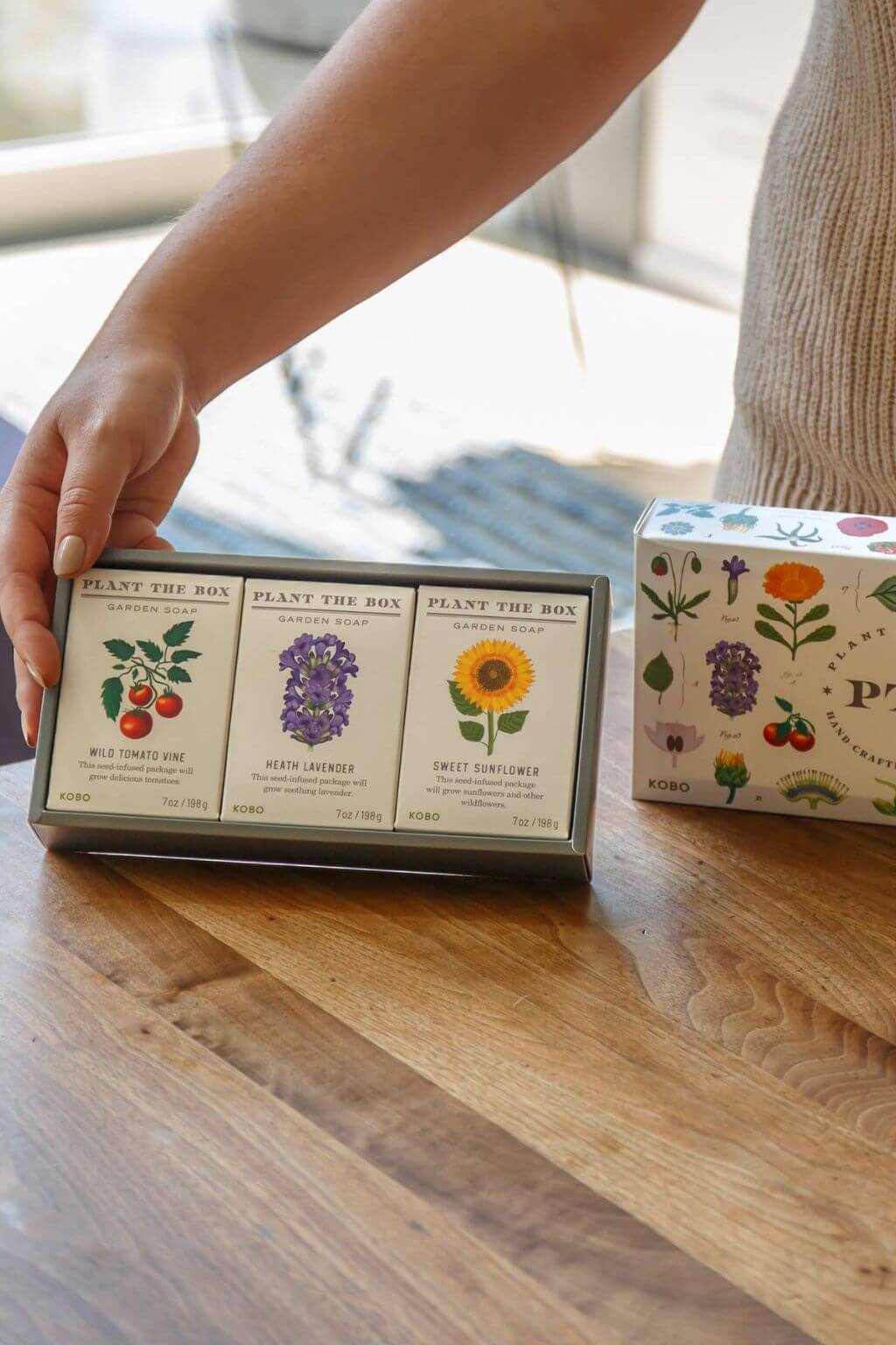 A person's hand holds a box of three Planty the Box scented soap bars on a wooden table, with another illustrated soap box visible in the background.