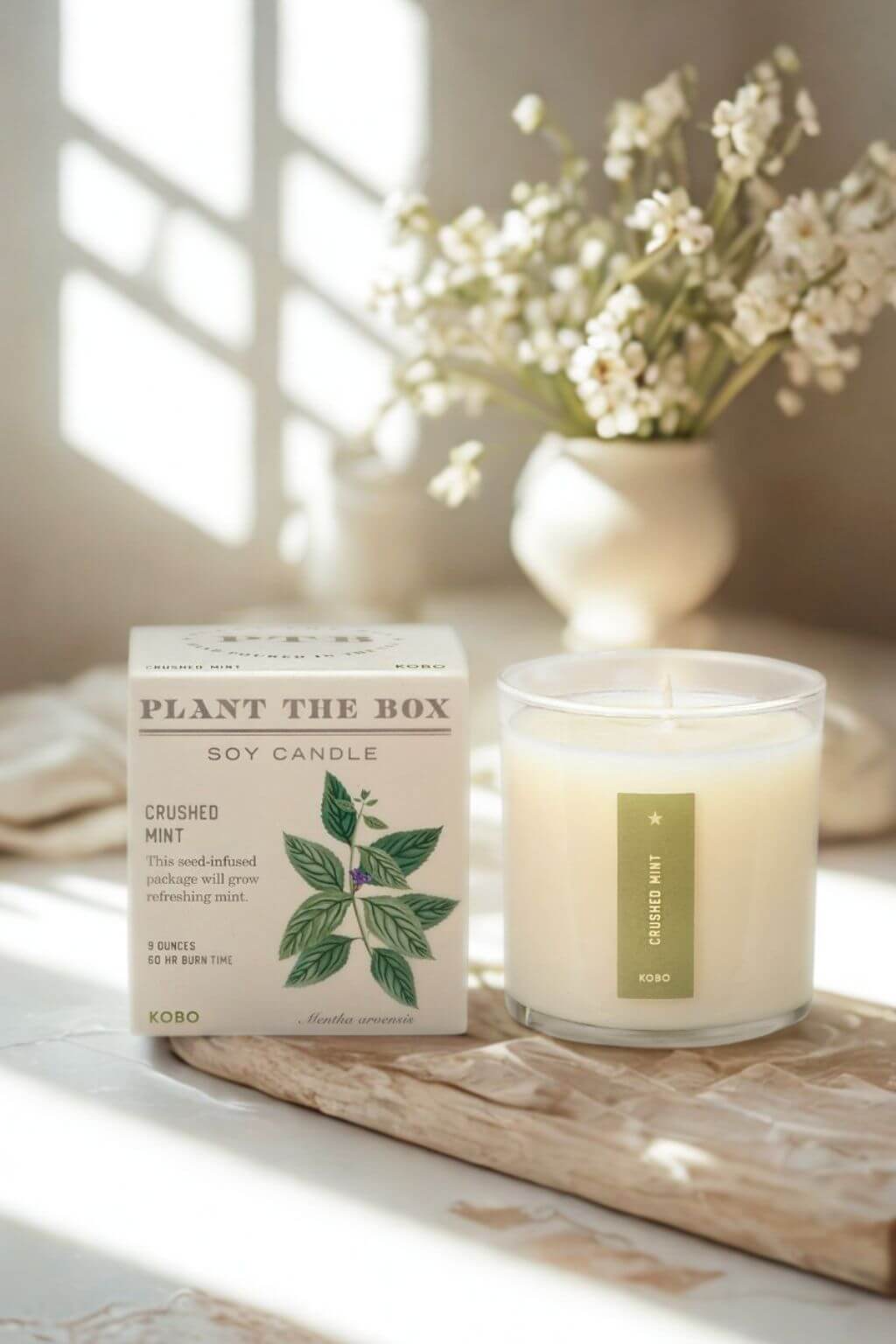 A soy candle in a clear glass jar sits beside its cream-colored product box on a wooden tray, with a blurred vase of white flowers in the background.