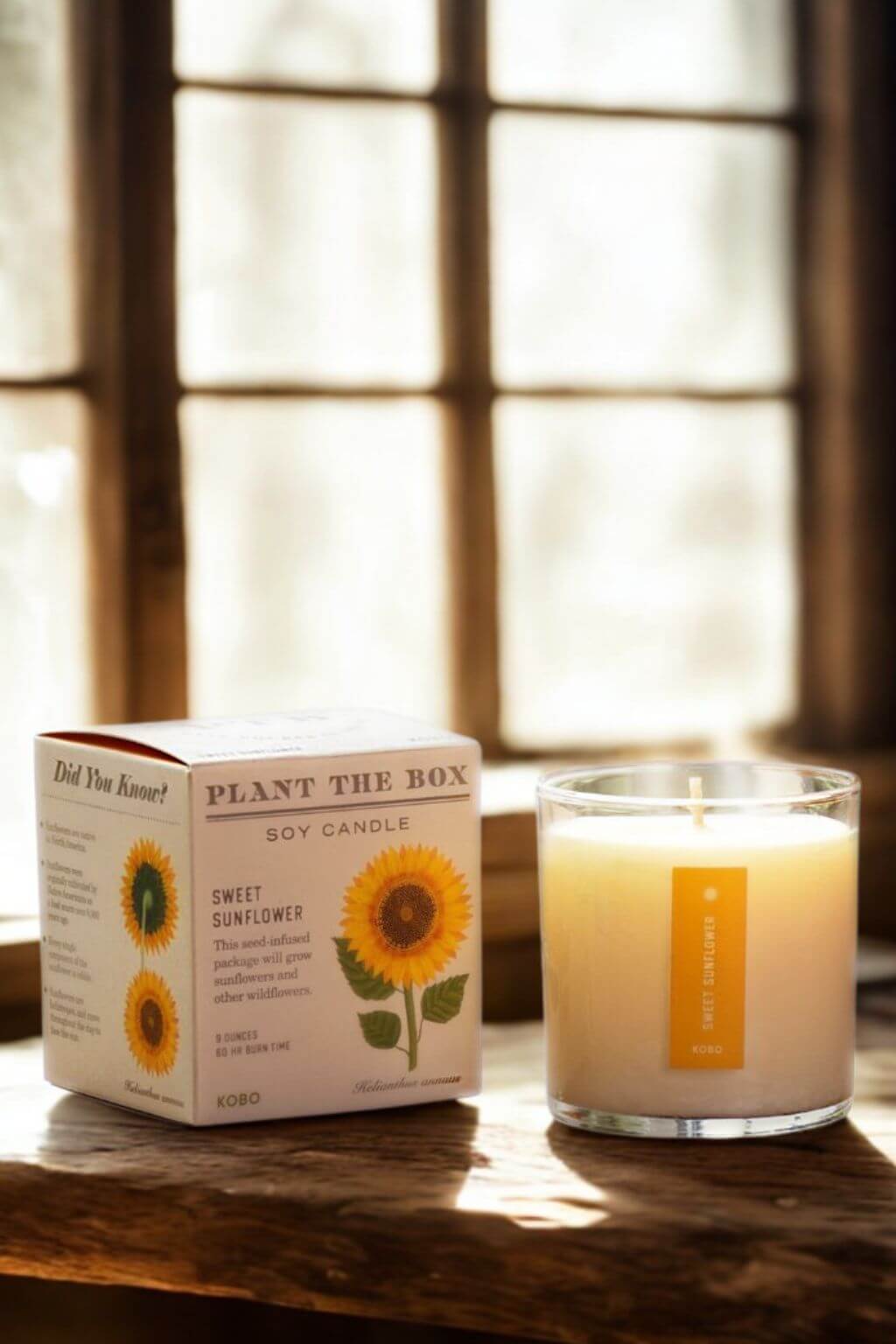 A lit soy candle in a clear glass jar sits on a wooden table beside its printed box. The packaging features a white background with yellow sunflower illustrations and the text 'PLANT THE BOX' and 'SWEET SUNFLOWER'.