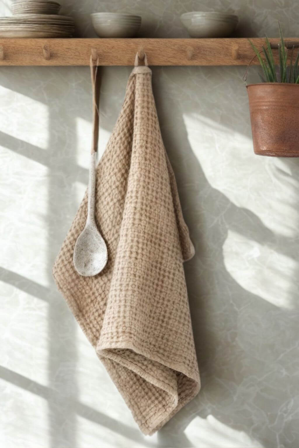 A beige waffle-weave kitchen towel hangs from a wooden shelf hook in a sunlit kitchen setting, with a white ceramic spoon resting in the center of the towel. The background features a textured light gray wall with soft window shadow patterns, a wooden shelf with stacked ceramic bowls, and a potted plant.