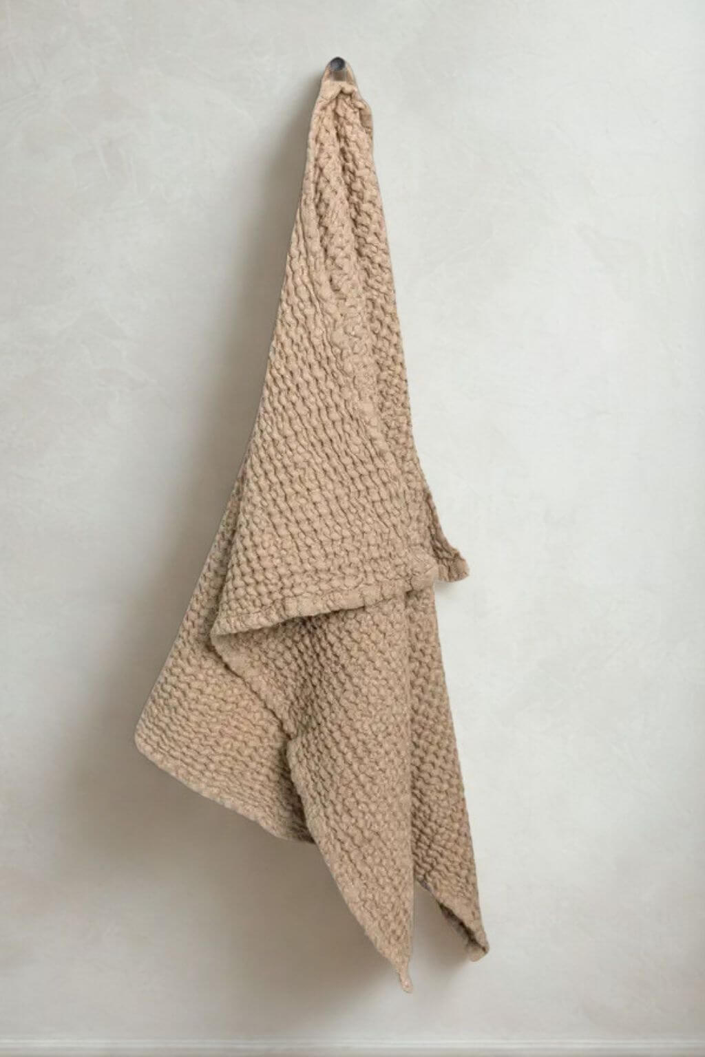 A chunky knit beige jhand towel draped over a wall hook against a textured off-white plaster wall.