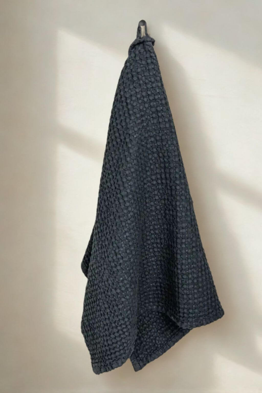 A dark slate blue textured waffle-weave kitchen towel hanging from a metal hook on a warm off-white wall with soft window light shadows.