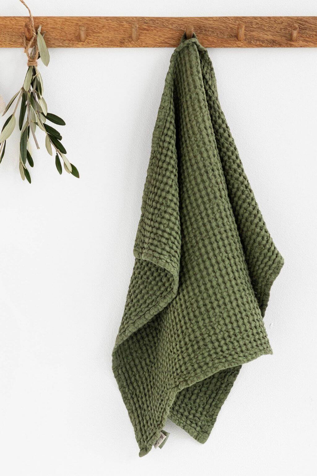 A forest green waffle-weave kitchen towel hanging from a wooden peg on a white wall, with a small olive branch decoration on the left.
