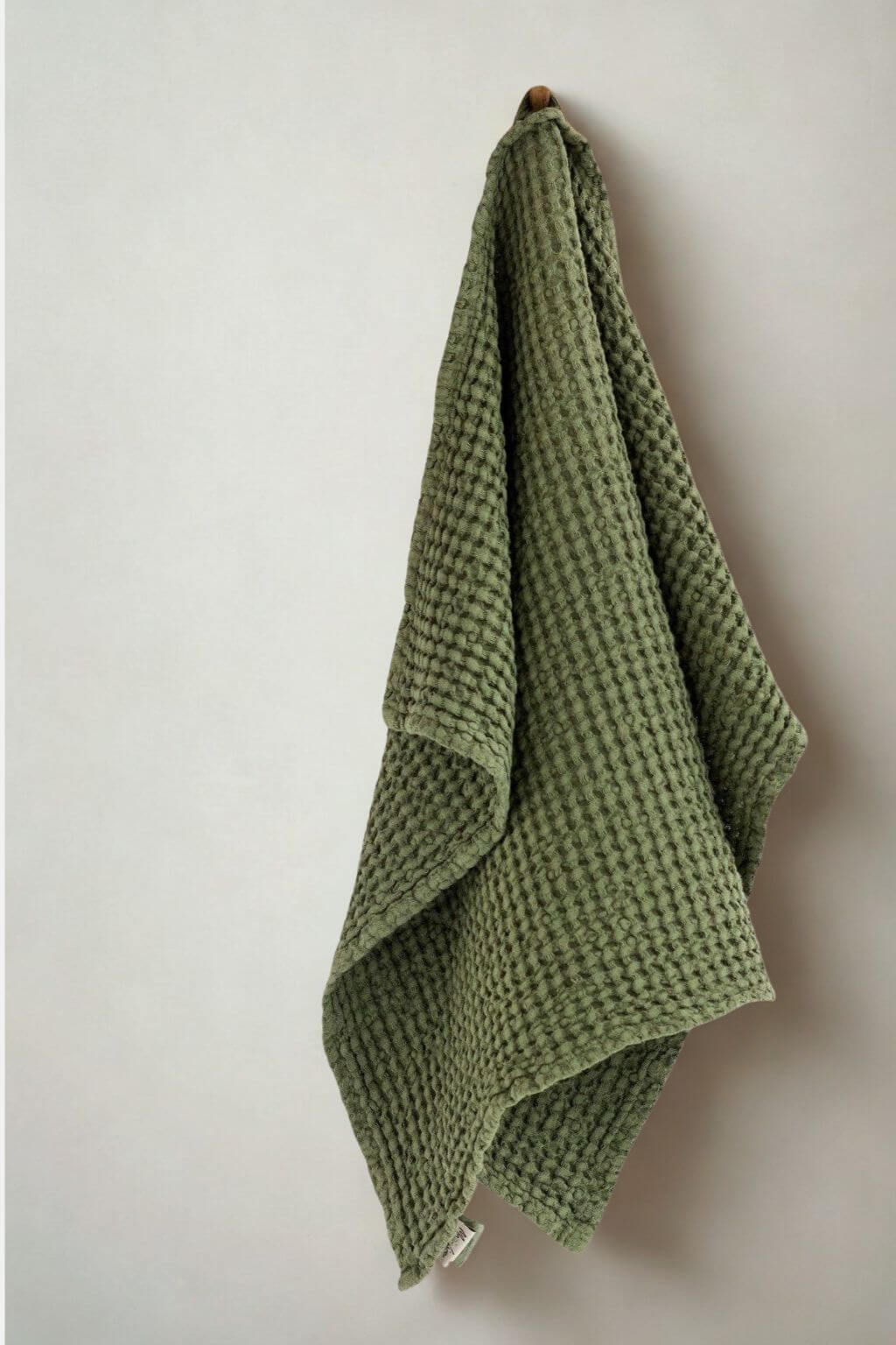 An forest green waffle-weave kitchen towel hanging from a small wooden hook against a plain off-white wall.