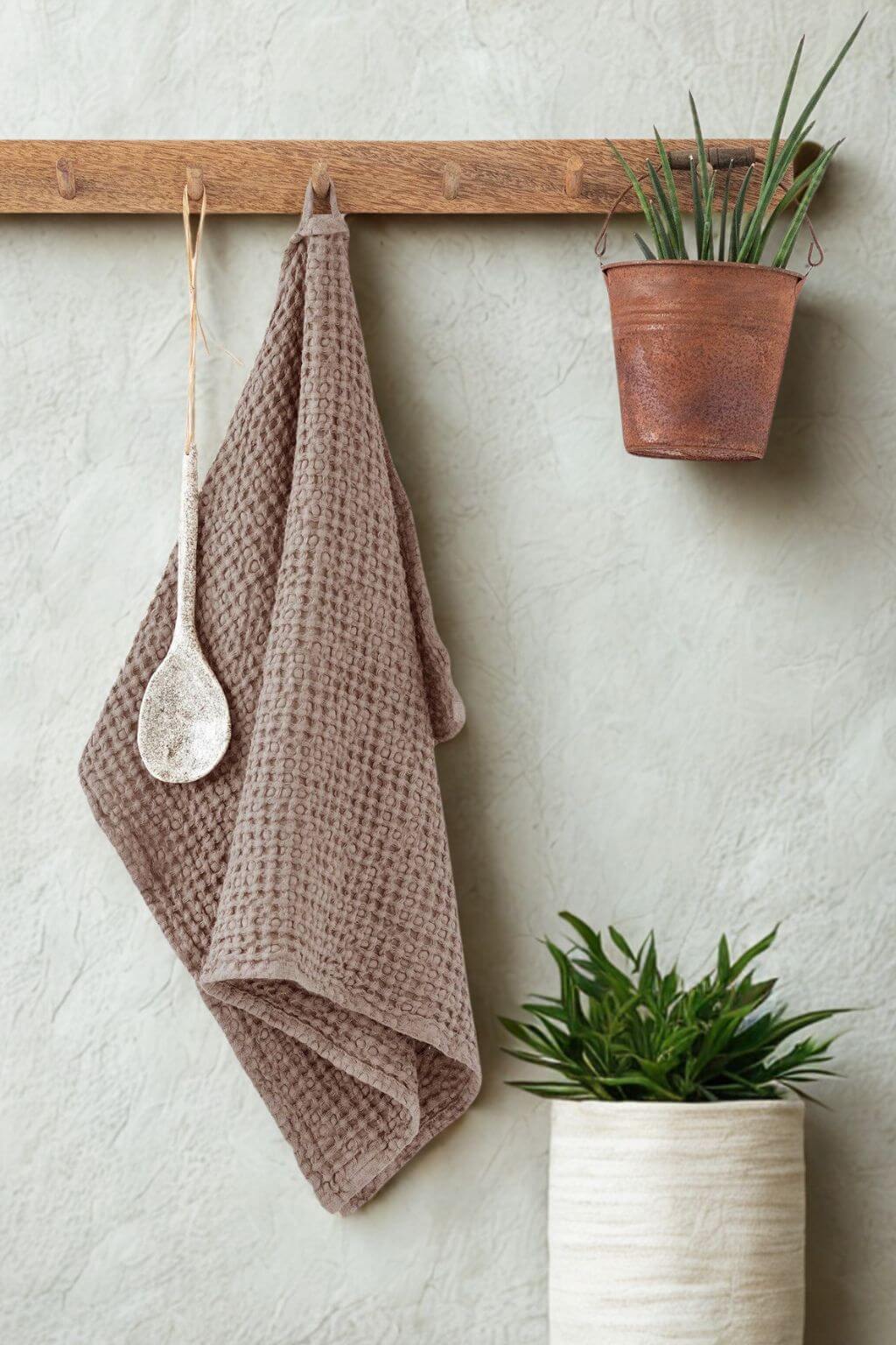 A textured waffle-weave kitchen towel in a warm taupe-brown color hangs from a rustic wooden peg rail on a textured plaster wall, alongside a wooden spoon and decorative potted plants.