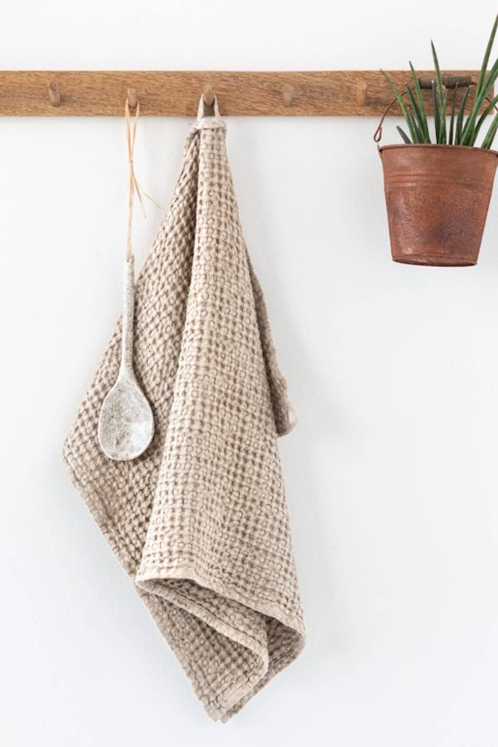 A sand textured waffle-weave kitchen towel hangs from a rustic wooden peg rail on a white wall, alongside a white ceramic spoon and a small potted green plant in a brown pot.