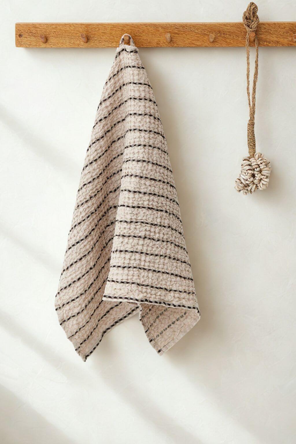 A textured beige kitchen towel with thin black stripes hangs from a wooden peg on a white wall, next to a hanging rope ornament.