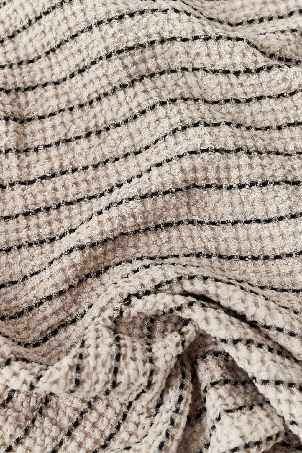 A close-up of a textured knit blanket with a waffle-weave pattern, featuring horizontal black stripes on a warm cream or beige background.