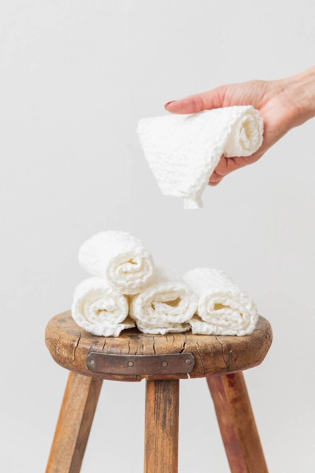 Hand holding a rolled-up washcloth above a stack of washcloths on a wooden stool