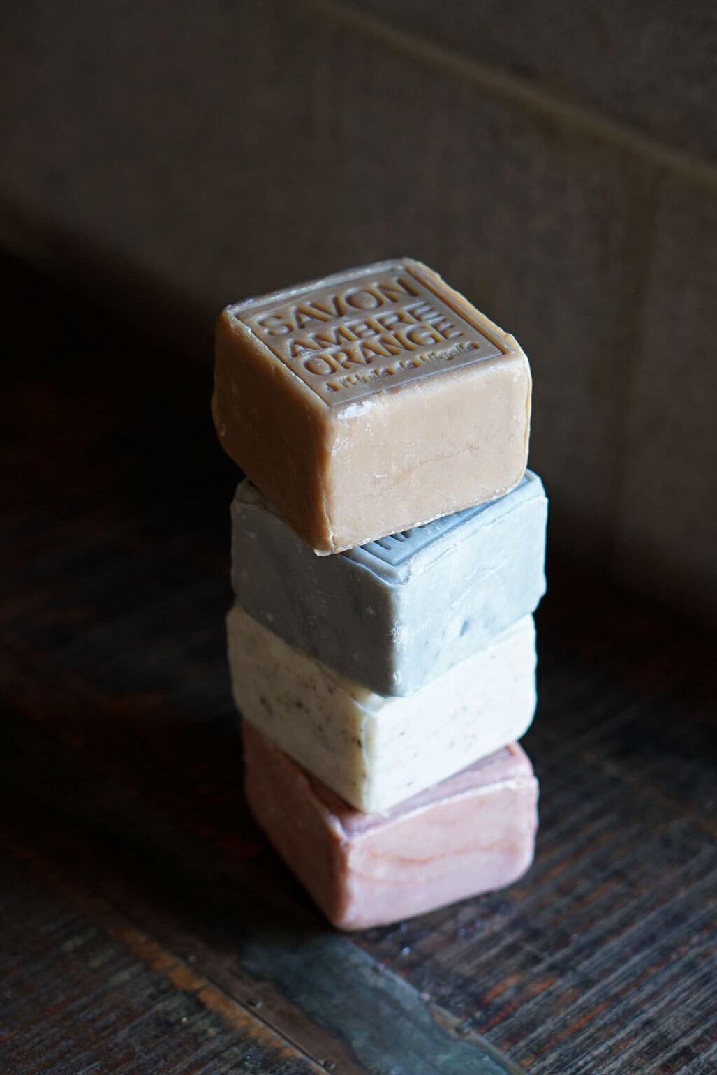Four stacked rectangular bars of soap in different muted colors, with the top bar embossed reading “SAPON AMBERE ORANGE,” photographed on a dark wooden surface against a dim indoor background.
