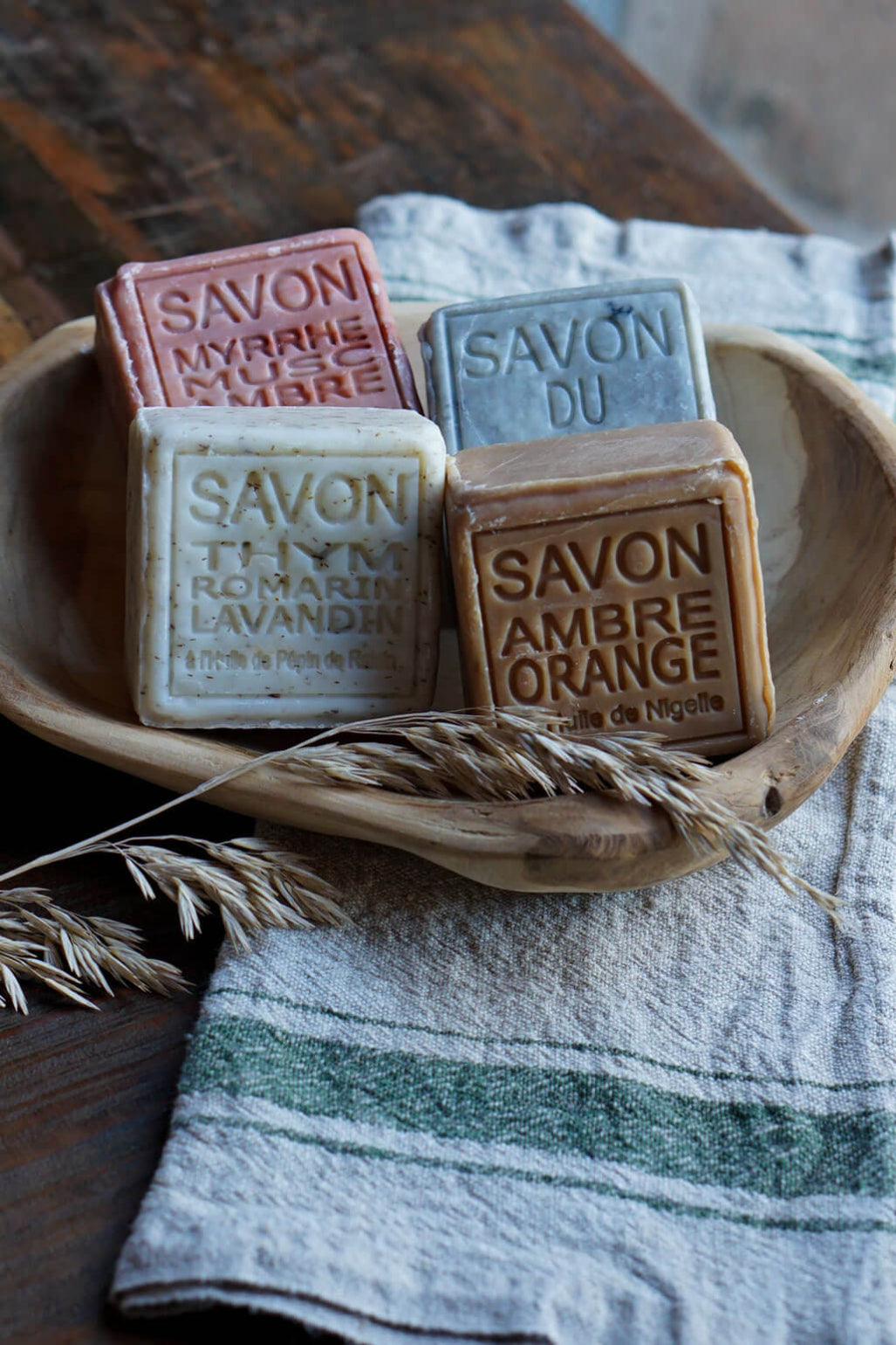 Four rectangular bars of artisanal soap in different colors, embossed with French text including 'SAVON MYRRHE', 'SAVON DU PUY', 'SAVON THYM ROMARIN LAVANDAN', and 'SAVON AMBRE ORANGE', arranged in a wooden bowl on a striped linen cloth with wheat stalks.