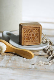 A rectangular bar of brown soap labeled 'SAVON AMBRE ORANGE' sits on a small white dish, with a wooden-handled brush in front and dried wheat stems on a rustic white surface.