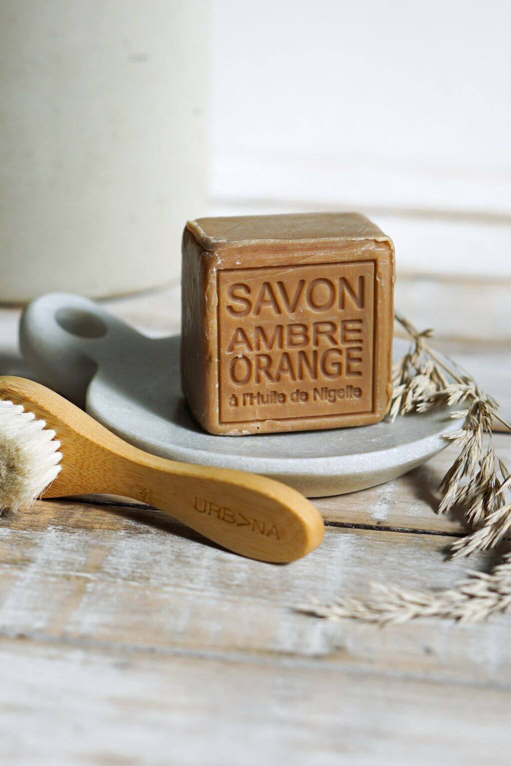 A rectangular bar of brown soap labeled 'SAVON AMBRE ORANGE' sits on a small white dish, with a wooden-handled brush in front and dried wheat stems on a rustic white surface.