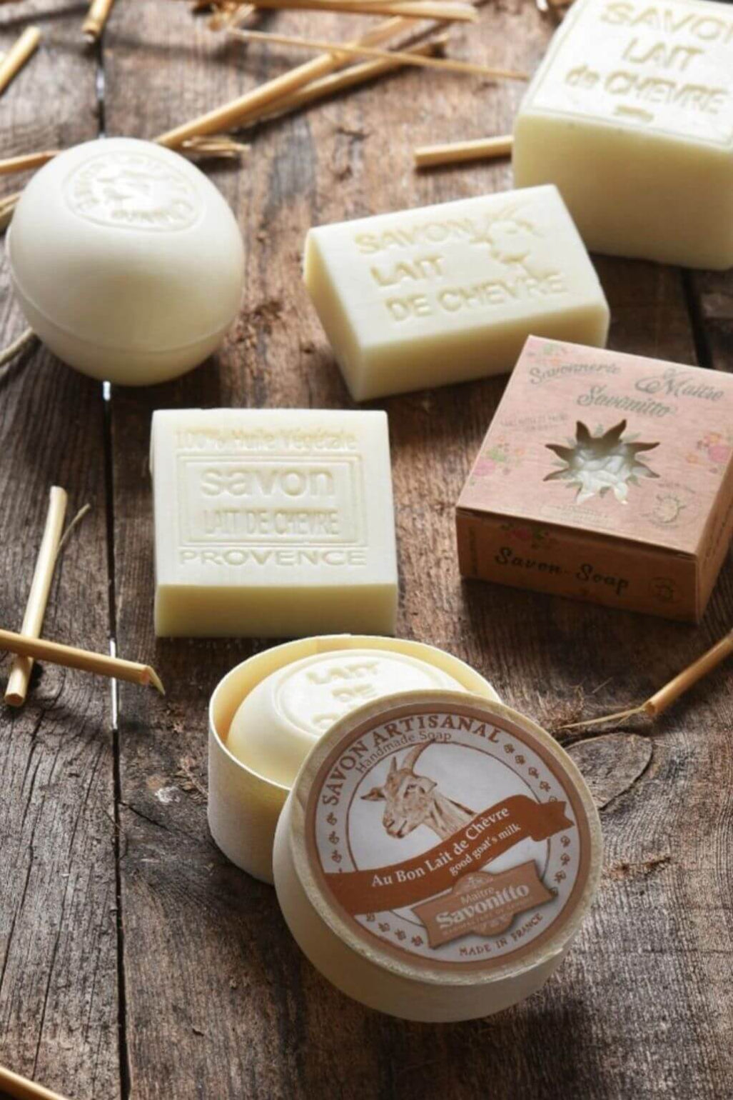 Several bars of handmade soap and a round tin of Savonitto soap are arranged on a rustic wooden surface with dried twigs.