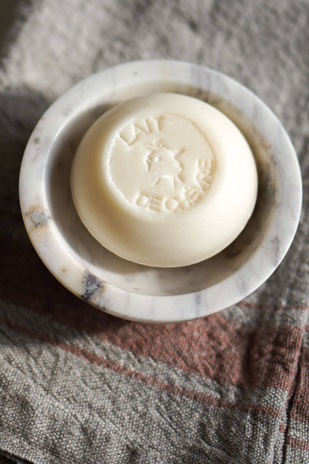 A round bar of Savonitto soap in an off-white cream color, resting inside a small white marble soap dish on a textured gray and brown linen cloth.