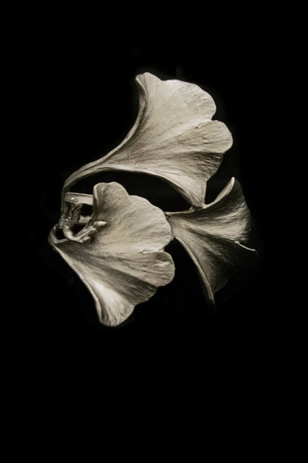 A silver napkin ring sculpted in the shape of three overlapping ginkgo leaves, shown against a solid black background.