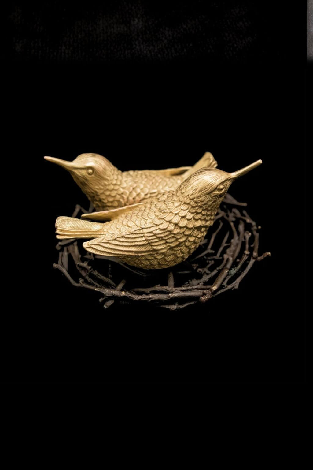 A small carved bird salt + pepper shakers in a warm gold tone, perched on a dark twig nest against a solid black background.