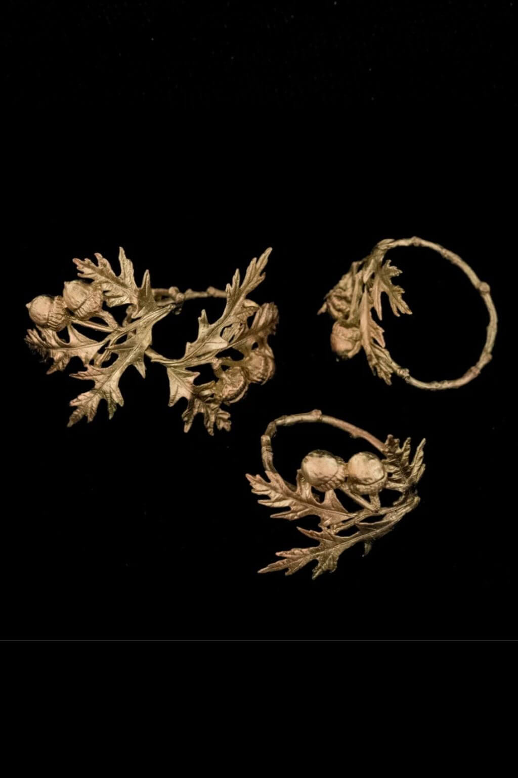 Four gold-toned vintage napkin rings with oak leaves and acorns, displayed on a solid black background.