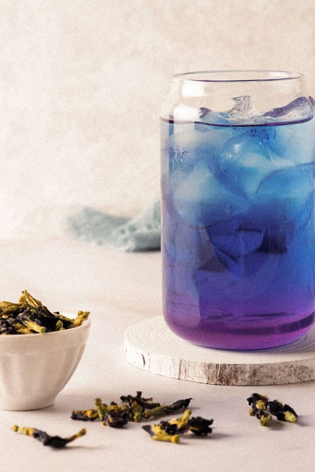 A clear glass can-shaped tumbler filled with a blue-to-purple iced drink and ice cubes, set on a round wooden coaster. A small white bowl with dried yellow-green flowers sits to the left, with a few scattered flowers on the surface.