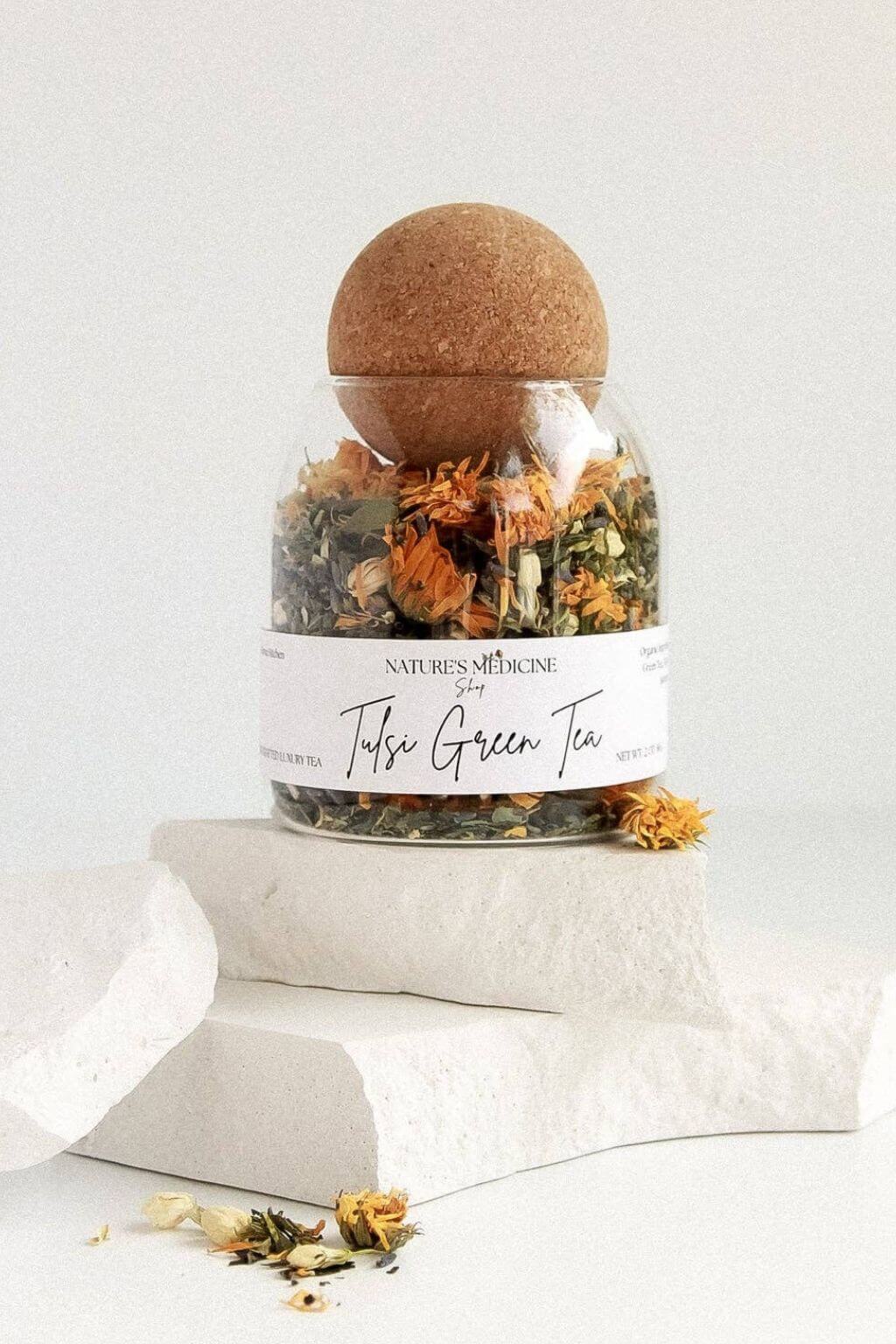 A clear glass jar of Nature's Muse Turmeric Green Tea with a cork stopper, displayed on white stone blocks against a clean white background.