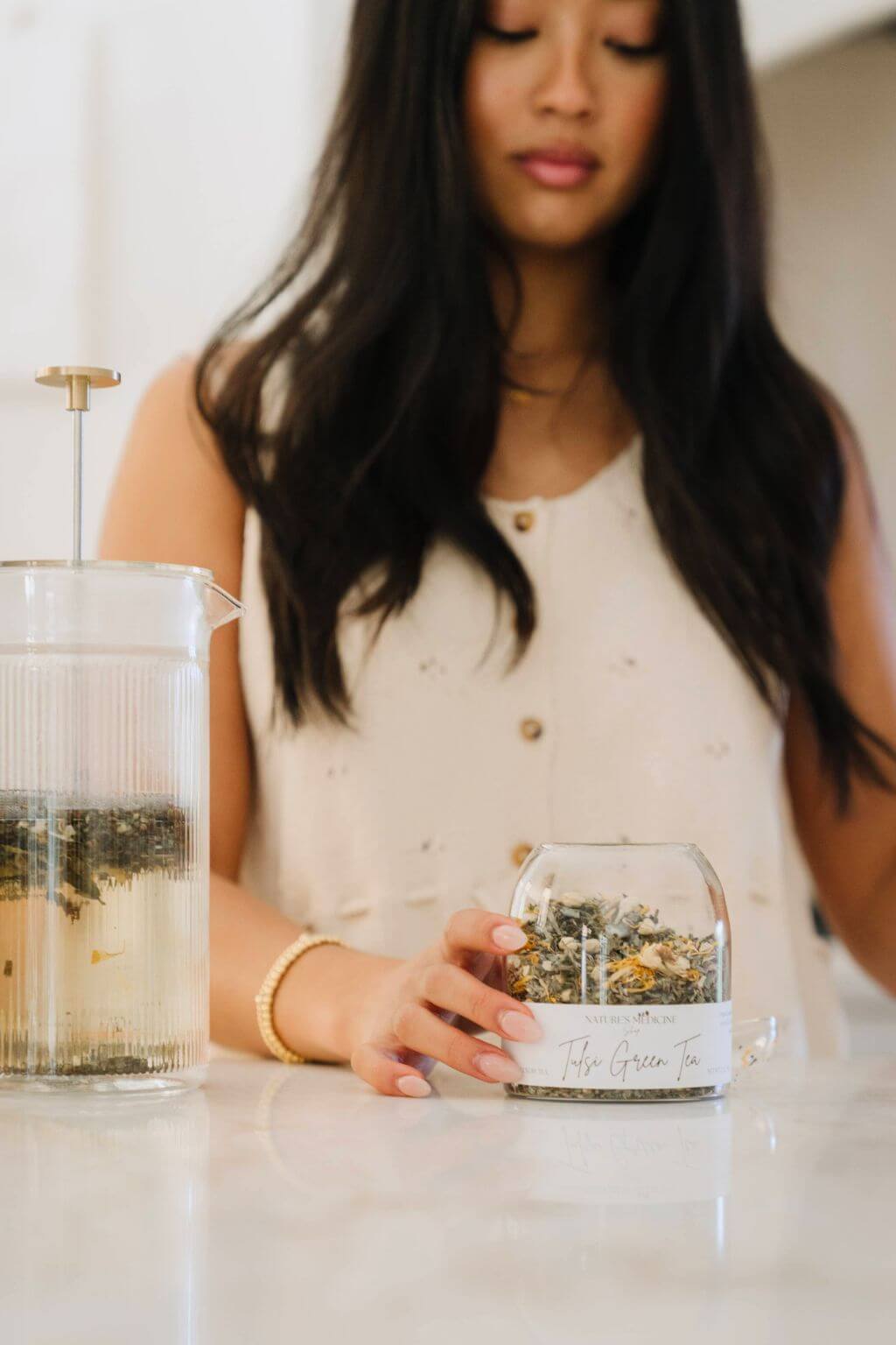 A woman in a white sleeveless top holds a small clear glass jar of loose-leaf herbal tea on a white countertop. A larger glass teapot with a metal plunger is visible to the left.