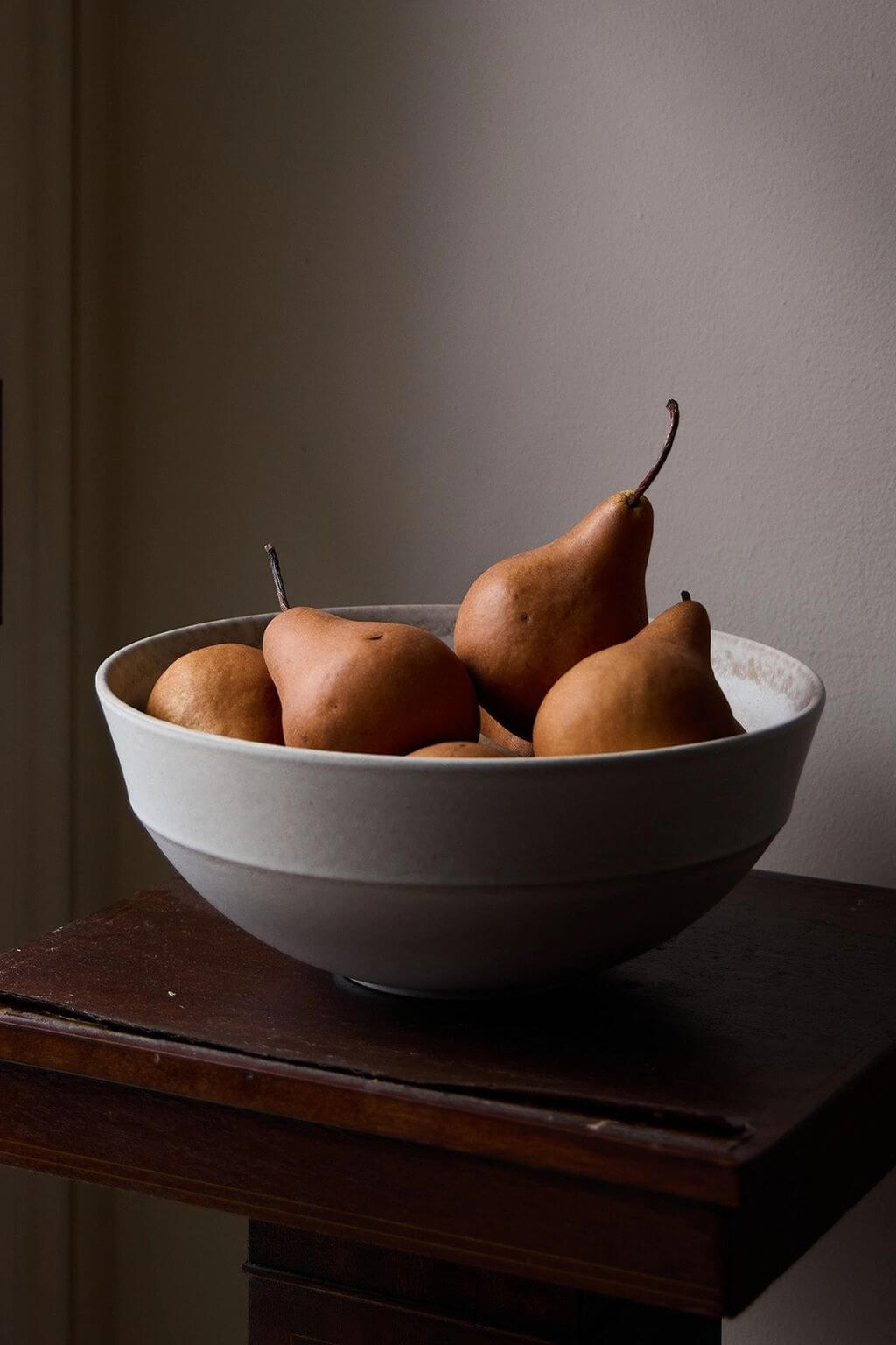 A white ceramic bowl filled with brown pears, set on a dark wooden table against a neutral beige wall.