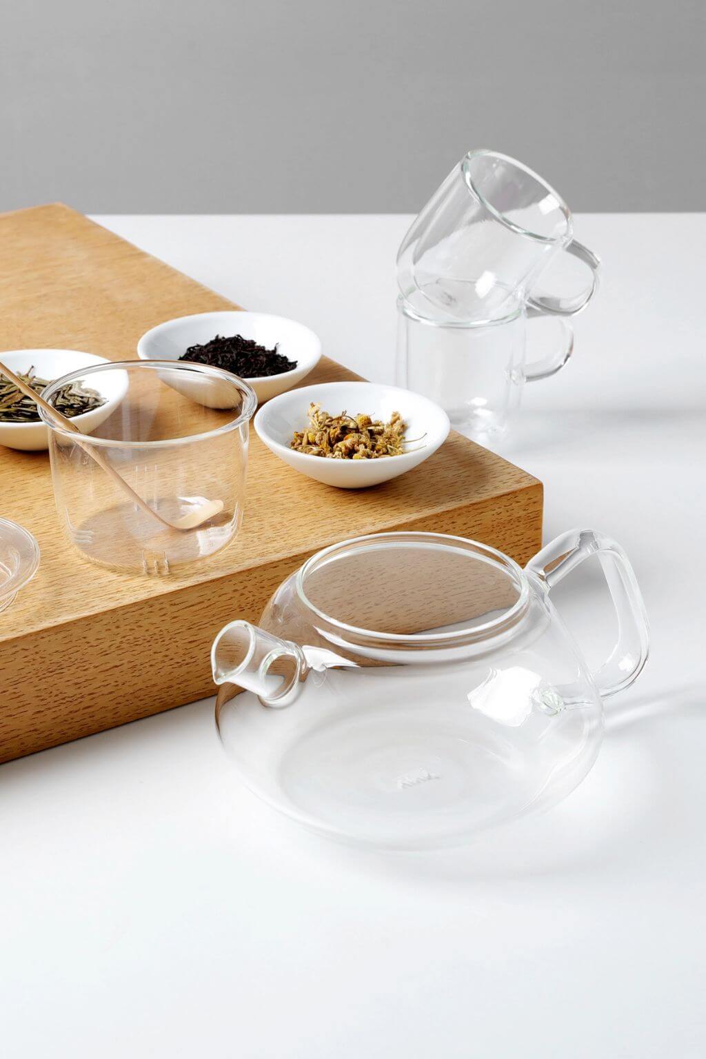 Clear glass teapot and cups on a wooden board with small bowls of tea leaves on a white surface.
