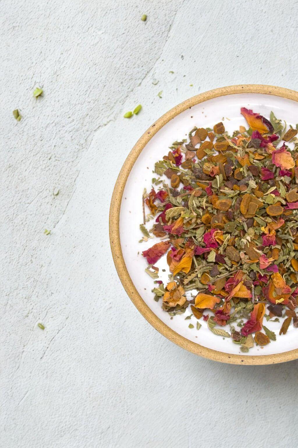 A top-down view of a white ceramic bowl with a natural clay rim, filled with loose-leaf herbal tea containing dried rose petals, green leaves, and orange flower pieces, set on a textured light gray surface.