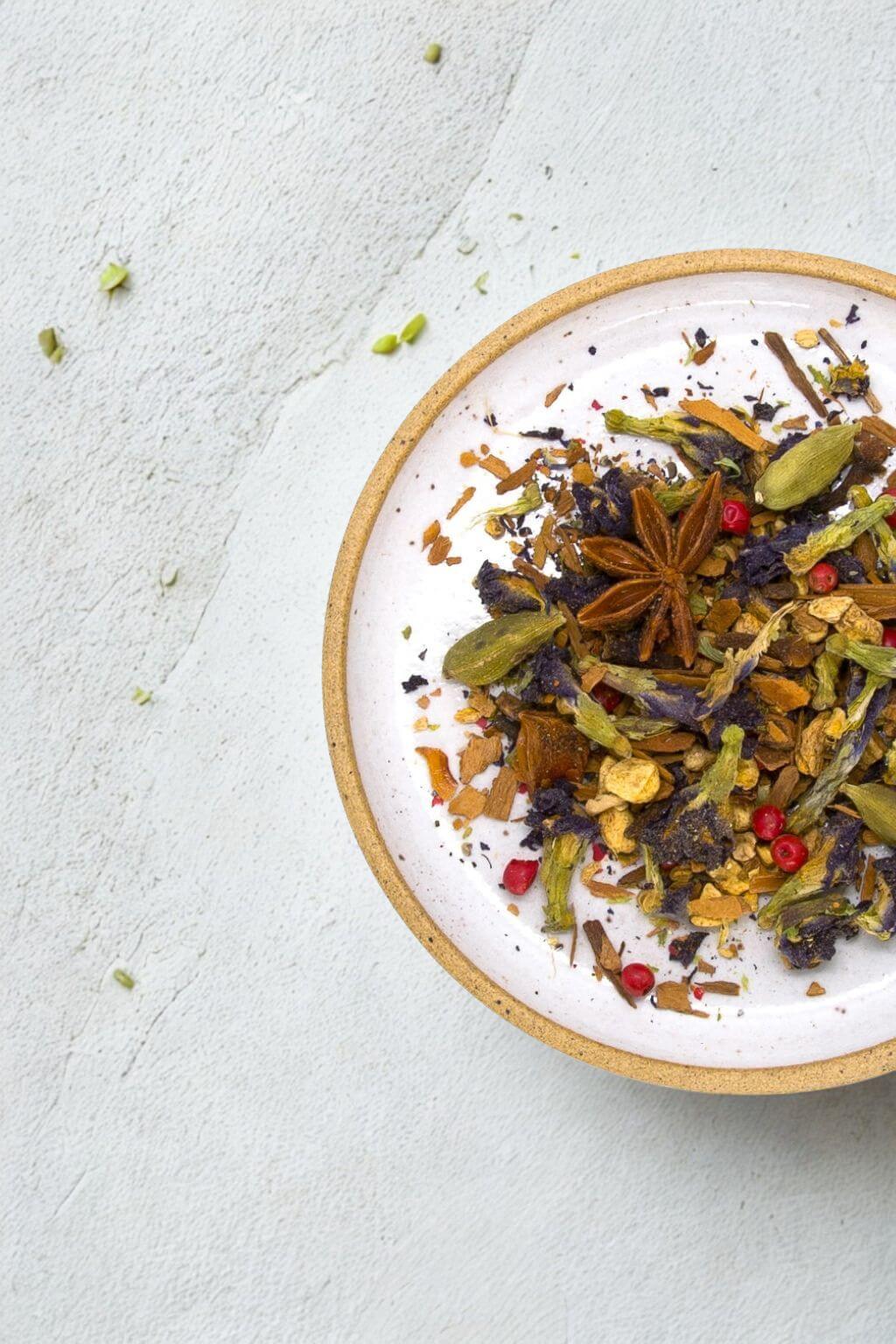 A top-down view of a white ceramic plate with a gold rim, filled with a colorful loose-leaf herbal tea blend containing star anise, green cardamom pods, dried flowers, and small red berries, set on a textured light gray surface.