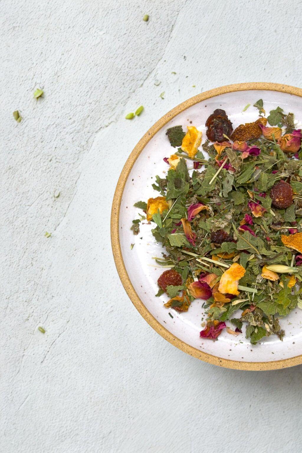 A ceramic plate with a natural clay rim filled with dried herbal tea blend, featuring green leaves, yellow flower petals, and dark berries, set on a textured light gray surface.