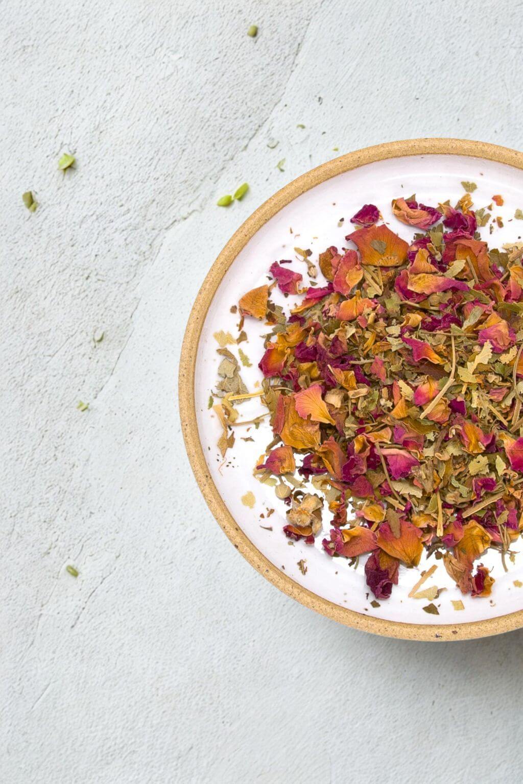 A top-down view of a white ceramic bowl with a natural clay edge, filled with dried rose petals and loose herbal tea, set on a textured light gray surface.