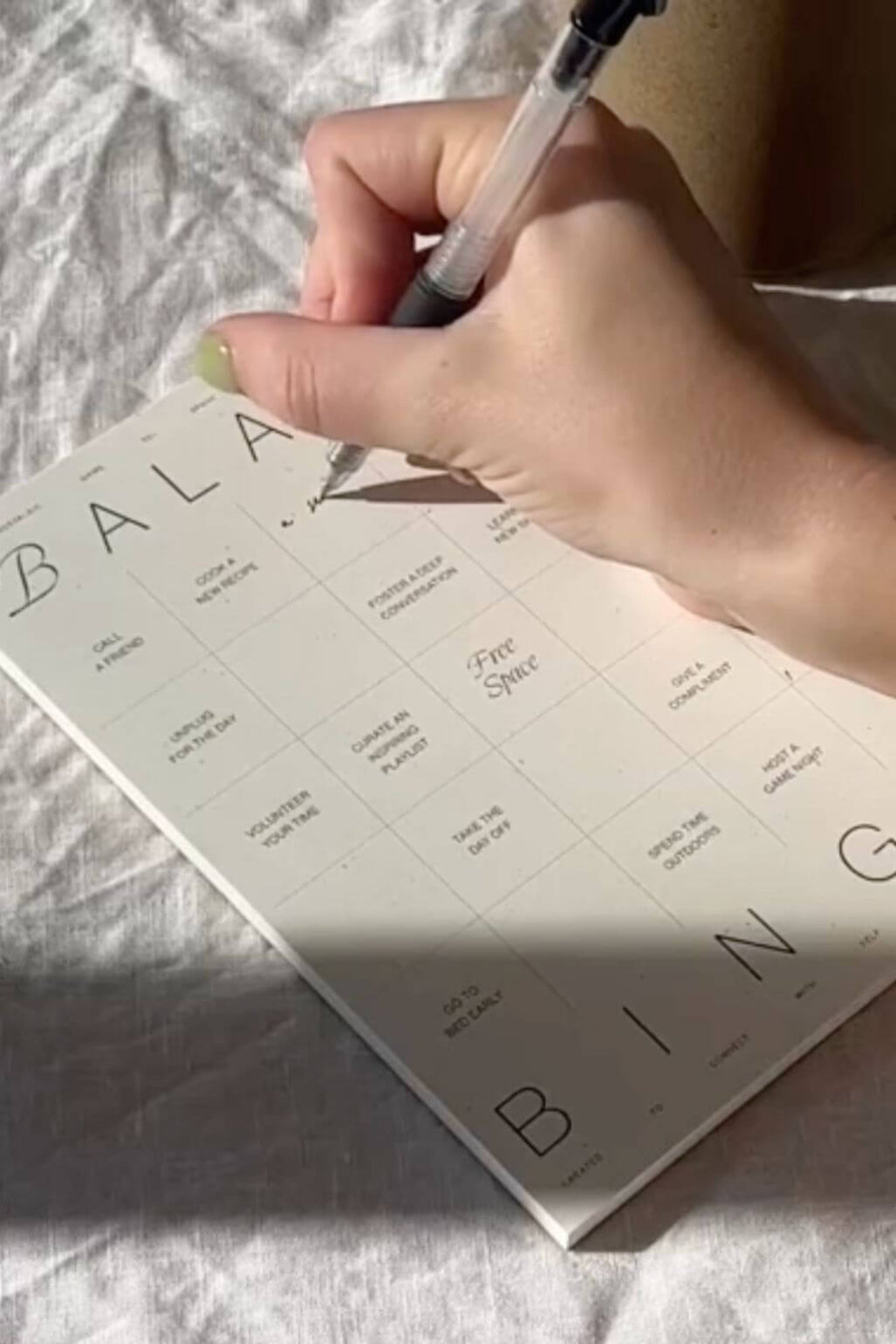 A close-up of a hand holding a black pen and writing on a white bingo card laid on a white fabric surface.